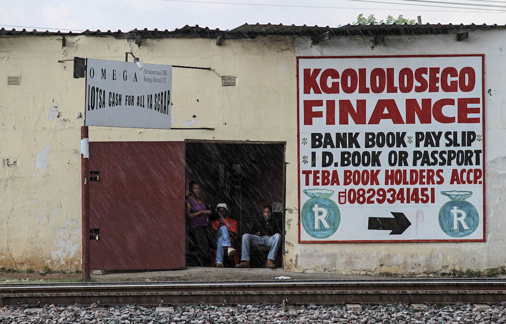 Residents wait inside a scrap metal merchant's office beside a money lender's advertisement for finance in Marikana.