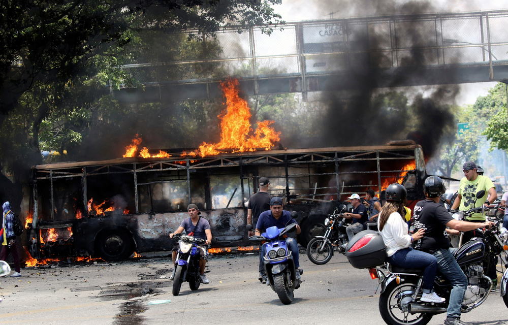 People drive their motorcycles near a burnt bus during anti-government protests