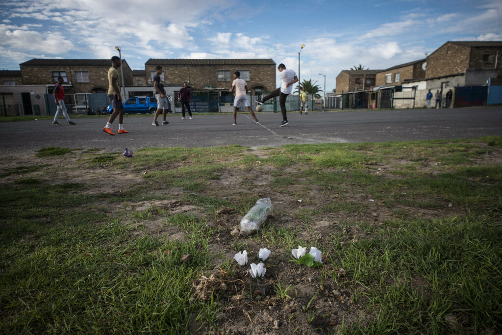 March 15 2022 Boys Play Football In Houw Hoek Park In Front Of What Is Left Of A Small White Flower Memorial To A Twenty Five Year Old Man Who Was Shot & Killed There On Wednesday 09 March. Gang Related Murders In Manenberg Over The Last Week Have Left